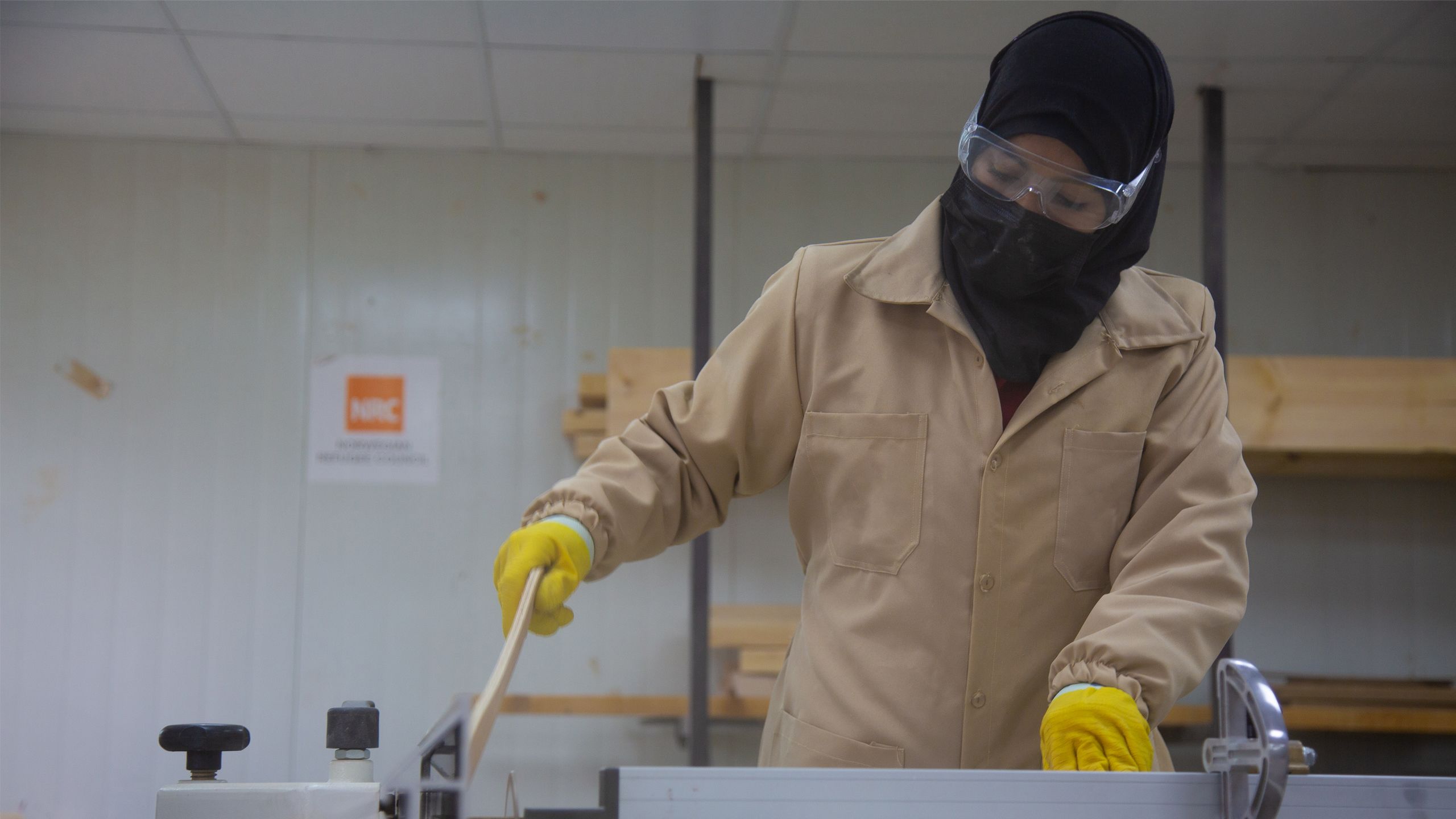 A woman is cutting a plank using a table saw in a wood workshop. She is is wearing protective goggles and yellow gloves.