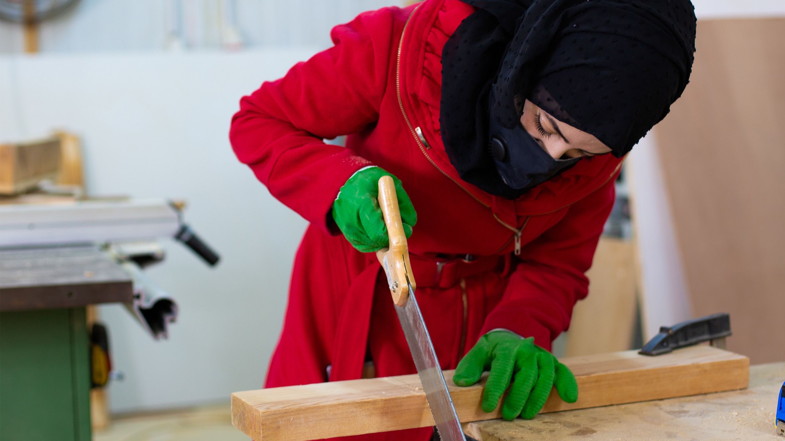 A woman in a red coat and green gloves is hand-sawing a wooden plank.