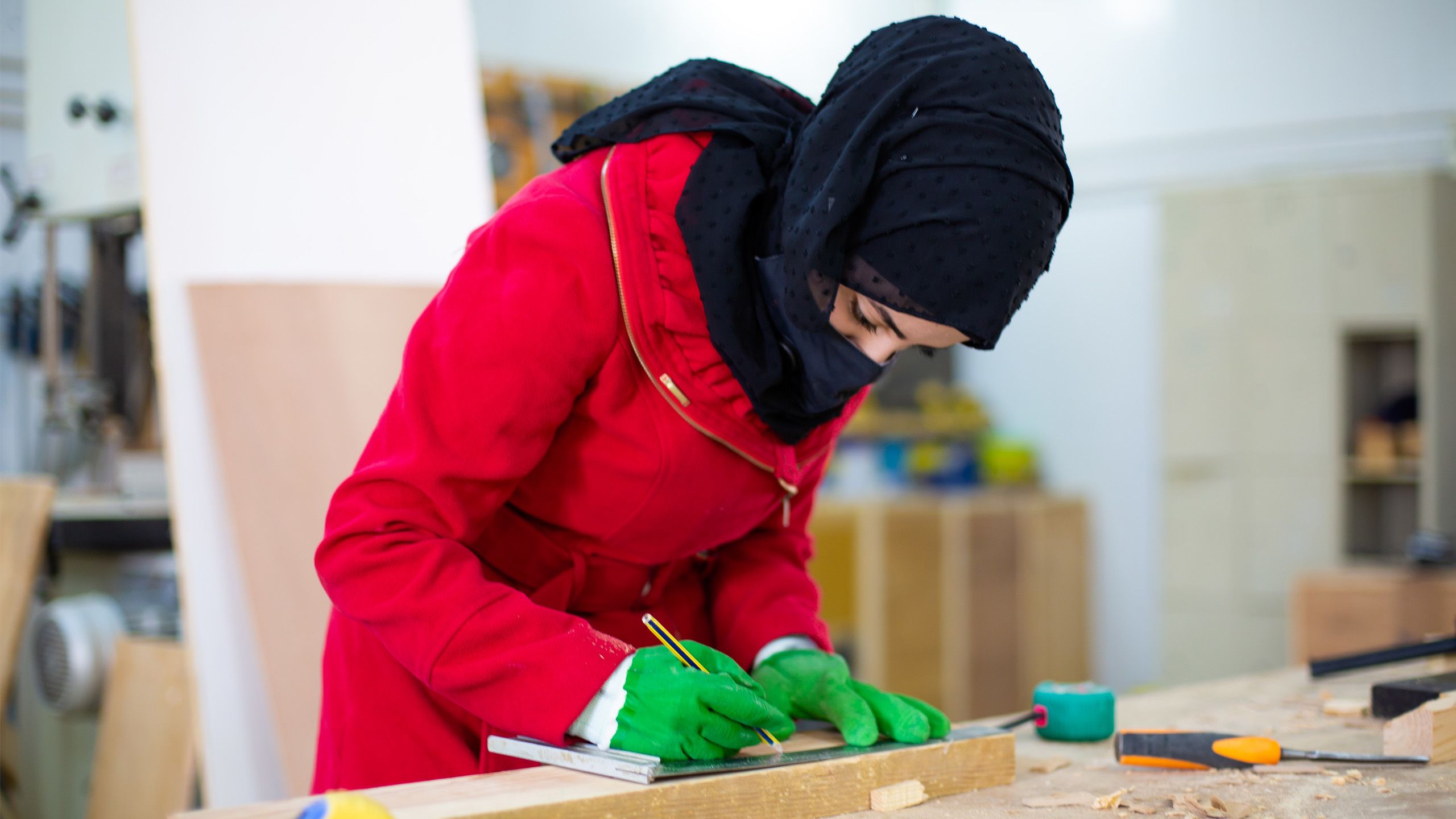 A woman in a red coat and green gloves marks the point at which a plank is to be cut. Around her is different carpentry tools strewn.