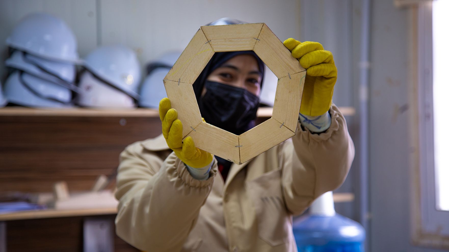 A woman in carpenter protective gear is holding up a wooden product she has made.